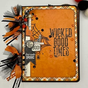 May include: A Halloween-themed scrapbook with an orange cover featuring the words "WICKED GOOD TIMES." The cover has a spiderweb design and the word "BOO!" is visible. The scrapbook is adorned with orange and black ribbons and embellishments.