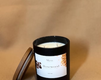 May include: A black candle with a wooden lid, labeled "Rosewood" by Mysa. The candle has a white label with brown accents and a brown wooden lid. The candle is on a brown surface.