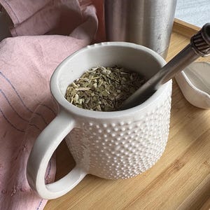 Puede incluir: Taza de cerámica blanca con superficie texturizada, llena de hojas de yerba mate secas. Una bombilla de plata está en la taza.