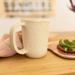 Puede incluir: Una taza de cerámica de color crema con una superficie exterior texturizada cubierta de pequeños puntos en relieve. La taza tiene un asa curva y resistente en el lateral y un borde liso y plano. El diseño es uniforme, con puntos por todo el cuerpo.
