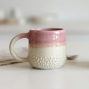 Puede incluir: Una taza de cerámica con un diseño de dos tonos, con una sección superior de color rosa suave y una sección inferior de color blanco crema. La mitad inferior de la taza está cubierta con un patrón texturizado de pequeños puntos en relieve.
