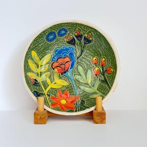 Puede incluir: Un plato de cerámica pintado a mano con un fondo verde y un diseño floral colorido. El plato presenta flores naranjas, azules y amarillas, con contornos blancos. El plato se exhibe sobre un soporte de madera.