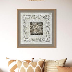 May include: A square wall art piece featuring a central dark woven panel surrounded by a white border with embossed spiral patterns and an outer layer of intricate white cut-out designs, all encased in a light brown wooden frame.