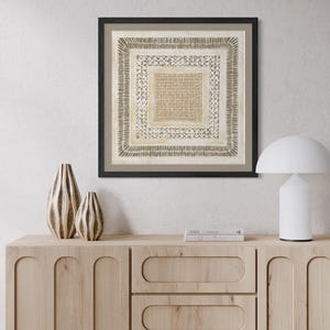 May include: A light wood sideboard with three drawers and two cabinets, topped with two textured brown vases, a stack of two books, and a white mushroom-shaped lamp. Above sits a square framed print featuring concentric patterns in beige and brown.