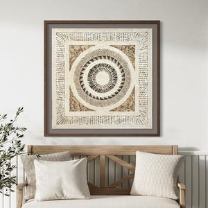 May include: A square framed wall art piece featuring a central circular mandala-style design with intricate black, white, and tan geometric patterns. The artwork is enclosed in a dark brown wooden frame and displayed above a light wood bench.