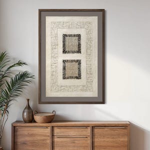 May include: A framed rectangular wall art piece featuring a cream-colored background with a border of wavy lines. The center contains two stacked square panels, each with a textured woven interior and a dark, fringed, spiky border design.