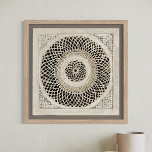 May include: A framed, square, off-white artwork with a circular, open-weave design. The artwork is set against a dark background and framed in a light wood. The piece is a decorative wall hanging, adding texture and visual interest to a space.