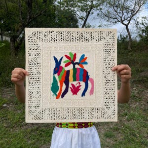 May include: A square, off-white textile with a colorful embroidered design. The central image features a vibrant donkey with a rainbow-colored body and a tree-like structure above it. The border has a cut-out geometric pattern.