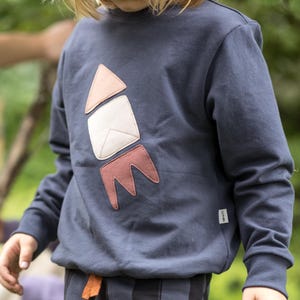 Könnte beinhalten: Marineblaues Sweatshirt mit einem Raketendesign in Rosa, Weiß und Braun. Das Sweatshirt wird mit einer gestreiften marineblauen Hose kombiniert. Das Outfit ist für Kinderkleidung geeignet.