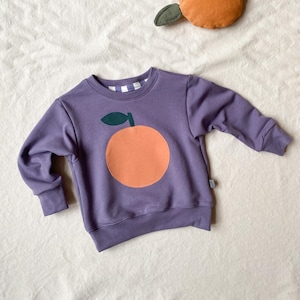 Könnte beinhalten: Ein lila Sweatshirt mit einem großen orangefarbenen Applikation und einem grünen Blatt. Das Sweatshirt hat lange Ärmel und einen Rundhalsausschnitt mit einem Karomuster im Inneren. Ein orangefarbenes Plüschtier mit einem grünen Blatt befindet sich im Hintergrund.