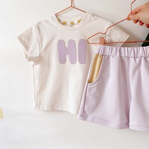 May include: A white short-sleeved t-shirt with a purple "HI" graphic and a pair of lavender shorts with a yellow pocket detail.