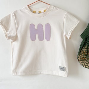 May include: A white short-sleeved t-shirt with a purple "HI" graphic on the front. The shirt has a crew neck and is made of a soft, comfortable fabric.