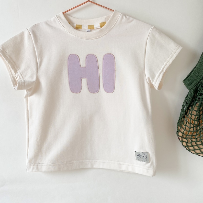 May include: A white short-sleeved t-shirt with a purple "HI" graphic on the front. The shirt has a crew neck and is made of a soft, comfortable fabric.