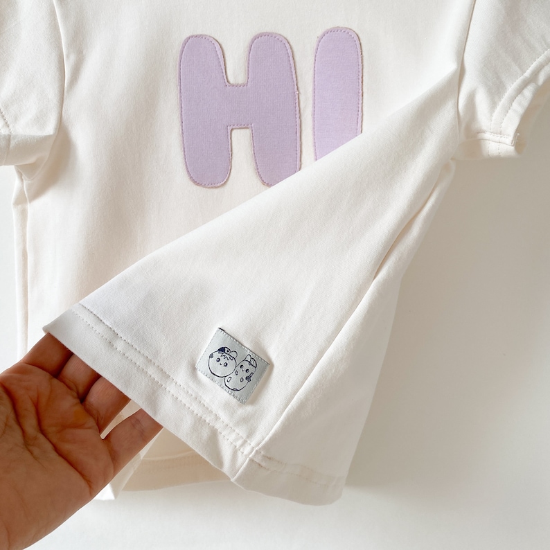 May include: A white t-shirt with a purple "HI" embroidered on the front. The shirt has a small tag with a cartoon design on the bottom.