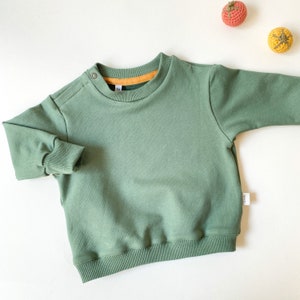 Könnte beinhalten: Salbeigrünes Sweatshirt mit geripptem Saum und Bündchen. Der Rundhalsausschnitt hat ein orangefarbenes Futter. Zwei kleine Knöpfe befinden sich auf der Schulter. Zwei gehäkelte Kugeln in Orange und Gelb sind im Hintergrund.
