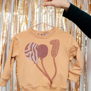 Könnte beinhalten: Ein hellbraunes Sweatshirt mit einem Ballon-Design. Die Ballons sind in Braun- und Lilatönen gehalten, einer mit Zebrastreifenmuster und ein weiterer mit weißen Punkten. Das Sweatshirt hängt an einem kupferfarbenen Kleiderbügel.