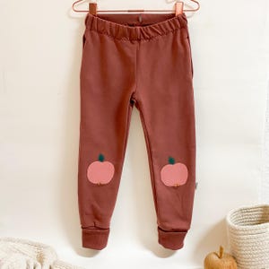 Könnte beinhalten: Rostfarbene Kinder-Sweatpants mit Apfel-Applikationen auf den Knien. Die Sweatpants haben einen Kordelzug in der Taille und gerippte Bündchen. Die Äpfel sind rosa mit grünen Blättern.