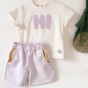 May include: A two-piece outfit for a child, featuring a white short-sleeved t-shirt with a purple "HI" graphic and lavender shorts with yellow side panels.