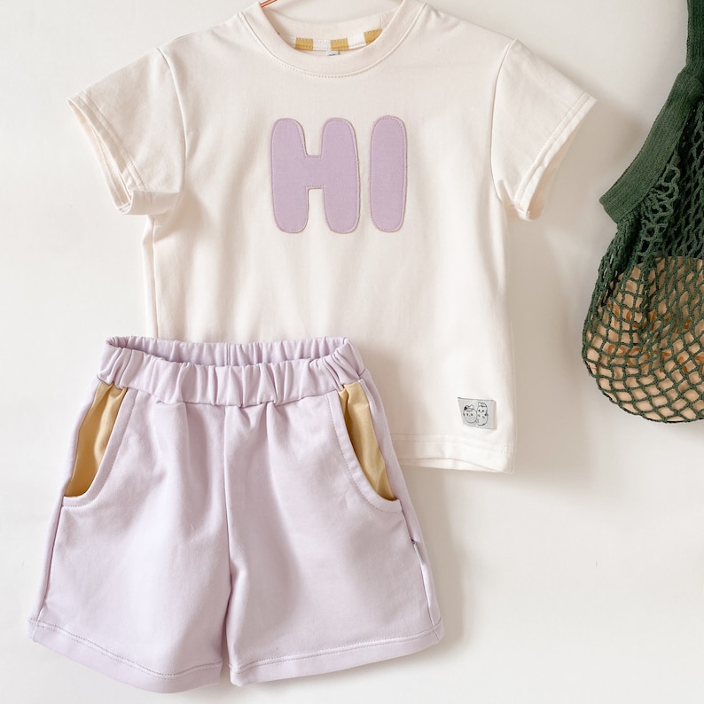 May include: A two-piece outfit for a child, featuring a white short-sleeved t-shirt with a purple "HI" graphic and lavender shorts with yellow side panels.