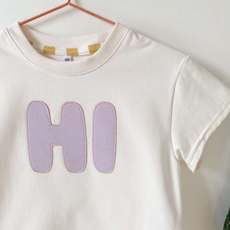 May include: A white t-shirt with a purple "HI" patch on the front. The shirt has a yellow and white striped trim around the neck.