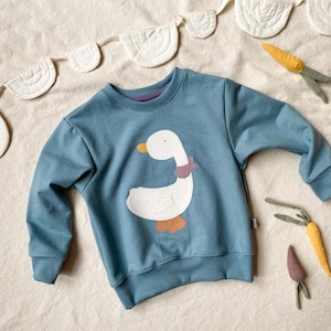 Könnte beinhalten: Ein blaues Sweatshirt mit einem weißen Gänse-Applikation, orangefarbenem Schnabel und Füßen sowie einem lila Schal. Das langärmlige Oberteil hat einen gerippten Saum und Bündchen. Dekorative Karotten und eine weiße Girlande sind ebenfalls zu sehen.