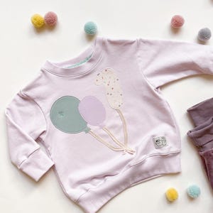 Könnte beinhalten: Ein lavendelfarbener Pullover mit einem Design aus drei Ballons, einem grünen, einem violetten und einem mit Konfetti-Muster. Der Pullover hat lange Ärmel und einen Rundhalsausschnitt.