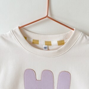 May include: A white t-shirt with a light purple applique of the letter "H" and a yellow and white striped trim around the neckline. The shirt has a size tag that says "98".