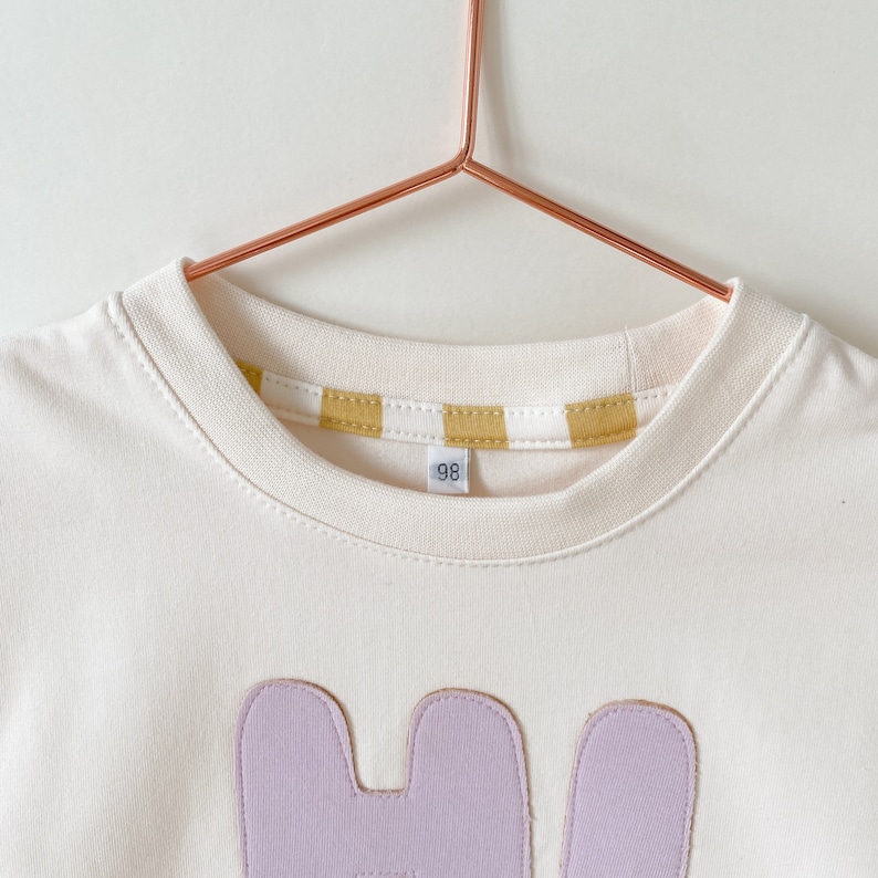 May include: A white t-shirt with a light purple applique of the letter "H" and a yellow and white striped trim around the neckline. The shirt has a size tag that says "98".