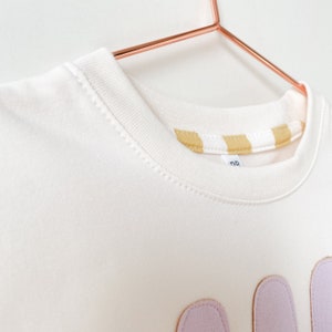 May include: White sweatshirt with a light purple applique of the word "Mom" on the front. The sweatshirt has a yellow and white striped trim around the neckline.