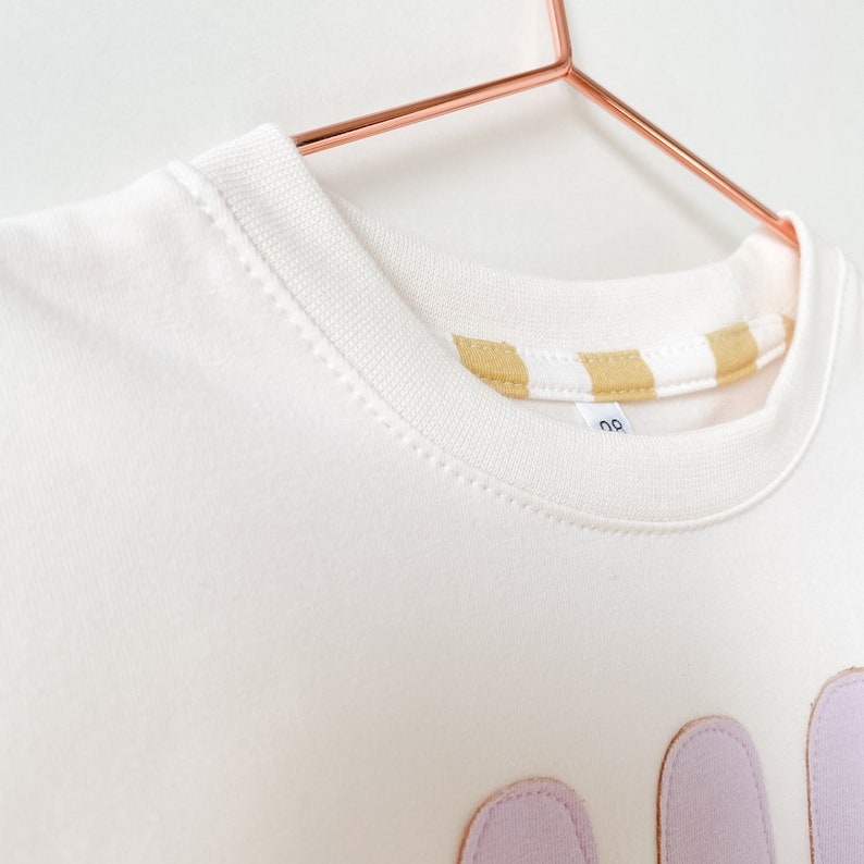 May include: White sweatshirt with a light purple applique of the word "Mom" on the front. The sweatshirt has a yellow and white striped trim around the neckline.