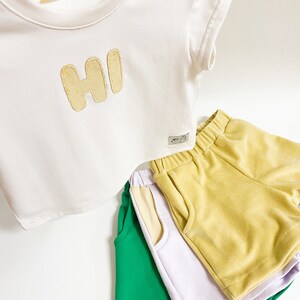 May include: A white short-sleeved t-shirt with the word "HI" in yellow felt letters. Three pairs of shorts in green, lavender, and yellow are stacked below the shirt.