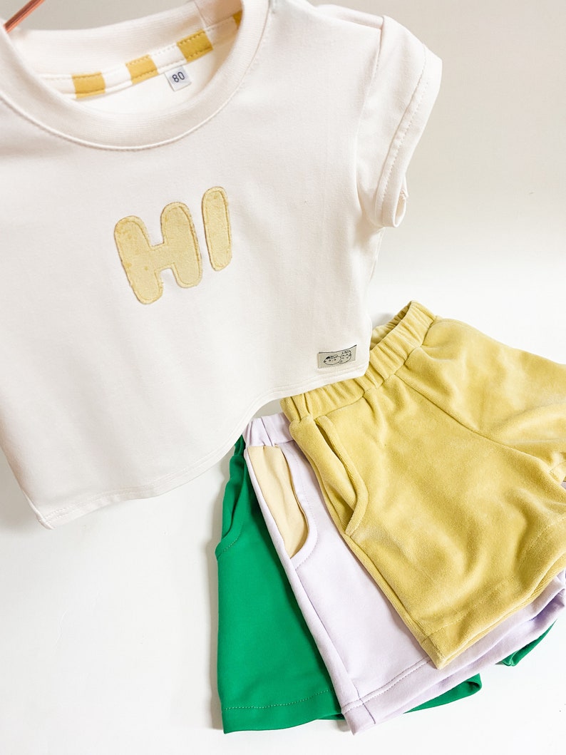 May include: A white short-sleeved t-shirt with the word "HI" in yellow felt letters. Three pairs of shorts in green, lavender, and yellow are stacked below the shirt.