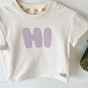 May include: A white short-sleeved t-shirt with a purple "HI" graphic on the front. The shirt has a yellow and white striped trim around the neckline.