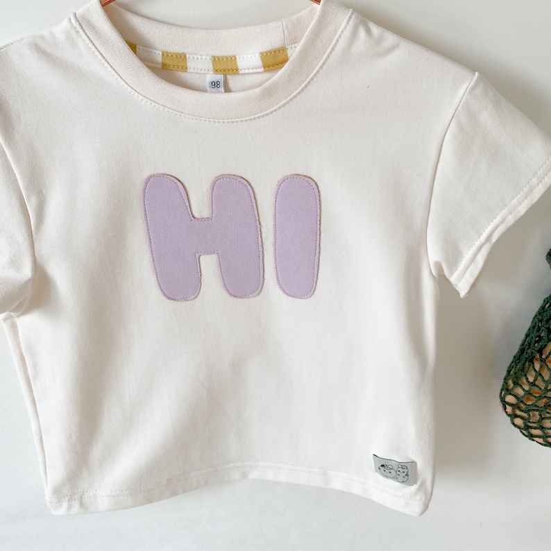May include: A white short-sleeved t-shirt with a purple "HI" graphic on the front. The shirt has a yellow and white striped trim around the neckline.