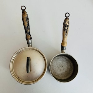 May include: Two vintage metal saucepans with wooden handles. One saucepan has a lid with a black handle. The other saucepan is open. The handles are a light brown color with black accents.