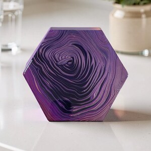 May include: A hexagonal coaster with a swirling, textured pattern in shades of purple and black. The coaster is made of a solid material and sits on a white surface. A vase of flowers is in the background.