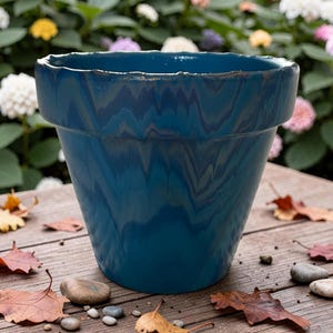 Peut inclure: Un pot de fleur en céramique bleu sarcelle avec un motif marbré ondulé. Le pot a un large bord et une forme conique. Il est posé sur une surface en bois avec des feuilles et de petits cailloux. En arrière-plan, des fleurs colorées floues.