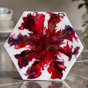 Puede incluir: Un posavasos hexagonal con fondo blanco y un vibrante diseño floral abstracto en tonos rojos, burdeos y morados. La obra de arte tiene un acabado brillante, creando un efecto visual llamativo.