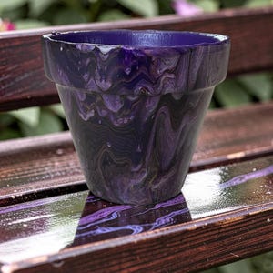 May include: A decorative flower pot with a marbled purple and black design. The pot has a traditional shape with a slightly flared rim. The colour scheme features deep purple and black hues, creating a unique, artistic appearance. Suitable for indoor or outdoor use.