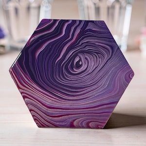 Pode incluir: Uma base de copo hexagonal com um design abstrato em espiral em tons de roxo, magenta e branco. A base de copo tem um acabamento brilhante e uma aparência artística única. É um item decorativo.