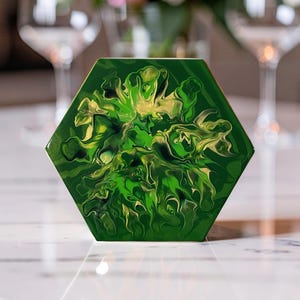 May include: A hexagonal coaster featuring a glossy finish with a marbled design in shades of forest green, lime green, and metallic gold. The surface displays fluid, swirling patterns that create a dynamic, organic appearance across the flat face.