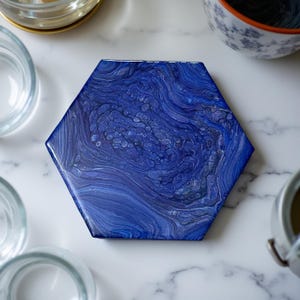 Könnte beinhalten: Sechseckiger Untersetzer mit einem Fluid-Art-Design in Blau- und Weißtönen. Der Untersetzer hat eine glänzende Oberfläche und ein einzigartiges, wirbelndes Muster. Ein dekorativer Artikel für die Wohnkultur.