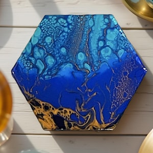 Könnte beinhalten: Ein sechseckiger Untersetzer aus blauer und goldener Kunstharz-Kunst mit einem wirbelnden, abstrakten Design. Der Untersetzer hat eine glänzende Oberfläche und ist perfekt, um Ihrem Zuhause einen Hauch von moderner Kunst zu verleihen.