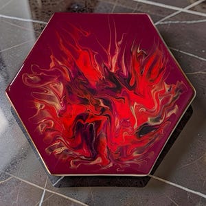 Könnte beinhalten: Sechseckiger Untersetzer mit burgunderfarbenem Hintergrund und einem lebendigen abstrakten Design in Rot, Schwarz und Gold. Die Kanten sind mit einem dünnen Goldrand versehen. Dieser Untersetzer ist für jede Einrichtung geeignet.
