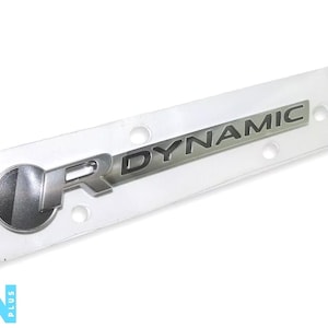 Puede incluir: Emblema plateado "R Dynamic" en un embalaje de plástico transparente. El emblema presenta las letras "R" y "DYNAMIC" en plata. Este emblema es para un coche.