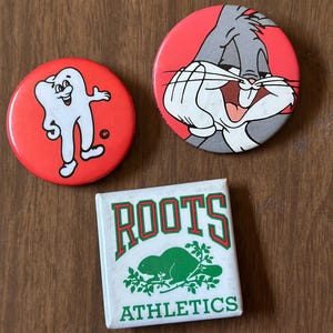 May include: Three round pinback buttons. The first button is red with a white cartoon tooth character. The second button is red with a cartoon Bugs Bunny character. The third button is white with green text that reads "ROOTS ATHLETICS" and a green beaver graphic.