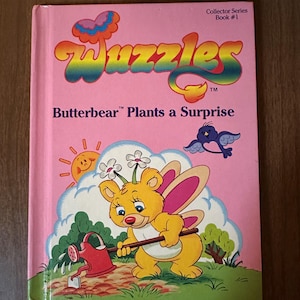 May include: A pink book with the title "Wuggles™ Butterbear™ Plants a Surprise" and a cartoon illustration of a yellow bear with pink butterfly wings watering a plant. The book is part of a collector series and is book #1.