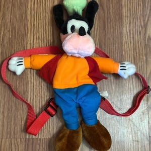 Może przedstawiać: Pluszowy plecak Goofy z zielonym czepkiem kucharza, pomarańczową koszulą, czerwoną kamizelką, niebieskimi dżinsami i brązowymi butami. Plecak ma czerwone paski.