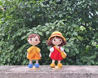 May include: Two crochet dolls standing side by side. The left doll has brown hair, a yellow dress, and blue shoes. The right doll has brown hair, a yellow hat, a white shirt, a red dress, and yellow shoes with a small yellow shoulder bag.