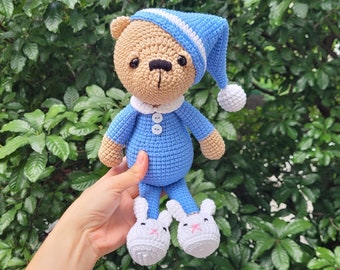 May include: A crocheted teddy bear wearing blue pajamas and a blue and white sleep cap. The bear is wearing white bunny slippers.